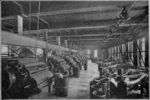 Fig. 122.   Wool carding. Notice the round cylinders. The gauzy web is being delivered into the can in front of the carding machine.