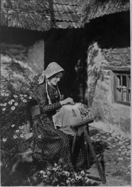 A maker of the Old Bucks Point lace at work, placing a pin into the parchment pattern. The picture shows also the hanging bunch of bobbins, and the old oak horse against which the lace pillow is rested.